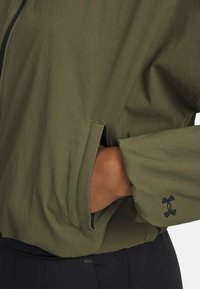Leichtgewichtige khakigrüne Jacke mit strukturierter Oberfläche, ausgestattet mit seitlichen Reißverschlusstaschen und einem schwarzen Logo auf dem Ärmel.