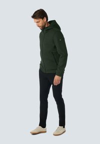 Groene zip-up hooded fleecetrui met capuchon, ribbelstructuur, zijzakken en logo-opdruk op de mouw, gecombineerd met zwarte broek en lichte sneakers.