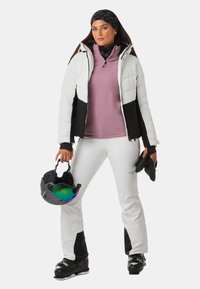 Skikleding met een wit en zwart pufferjack, een roze fleece top, witte broek en zwarte skibotten. Houdt een helm vast met een getinte vizier.