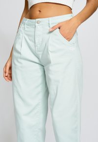 Světle mátově zelené kalhoty s vysokým pasem a volnějším střihem, vybavené předními kapsami, zapínáním na knoflíky a malou značkovou nášivkou na kapse.