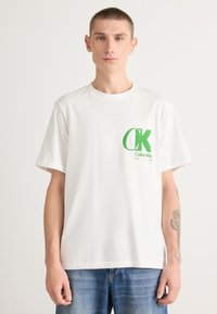 Camiseta de algodón blanca con un logo verde de "CK" en el lado izquierdo del pecho. Mangas cortas, escote redondo, corte relajado y textura suave.