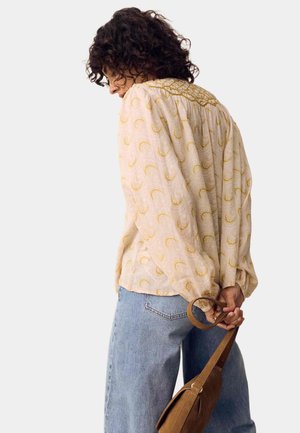 Persoon met krullend donker haar, een beige blouse met patroon en een blauwe spijkerbroek, die een bruine tas bij de band achter hun rug vasthoudt.