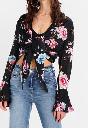 Kvinde iført sort blomsterprintet langærmet bluse bundet i taljen, kombineret med højttaljede blå jeans og en guldfarvet vedhængshalskæde.