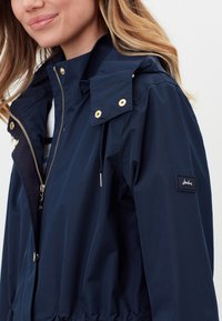 Navy sininen vedenpitävä takki, jossa on vetoketjusuljin, kultasävyiset yksityiskohdat, nyörikiinnitys vyötäröllä ja brändin logo vasemmassa hihassa.