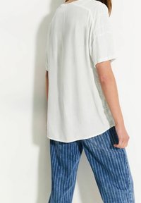 Vit kortärmad skjorta, lätt material, avslappnad passform, rundad nederkant, bärs med blå vertikalt randiga byxor. Minimalistisk design och textur.