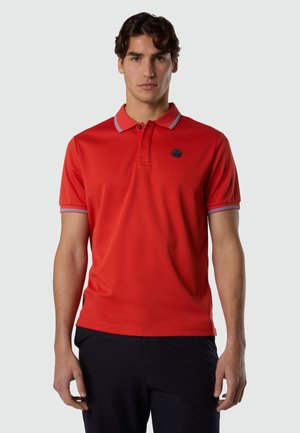 Mann trägt ein rotes Kurzarm-Poloshirt mit grauer Einfassung an Kragen und Ärmeln, steht vor einem schlichten hellen Hintergrund.