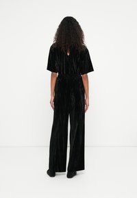 Combinaison en velours noir à manches courtes, jambes larges et élastique à la taille. Comprend un détail en trou de serrure dans le dos. Texture lisse.