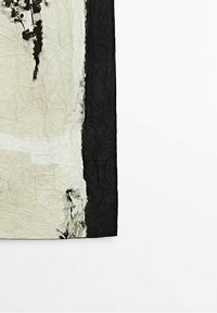 Tissu froissé avec une bordure noire verticale sur le côté droit et des marques noires abstraites sur un fond beige.