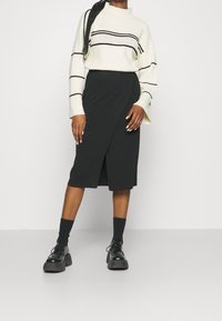 Gräddfärgad ribbad tröja med svarta ränder i kombination med en mörkgrå omlottskirt. Accessoarer inkluderar svarta snörskor och ankellånga strumpor.