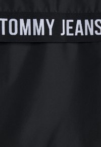 Zwart materiaal met een gladde textuur, met een opvallend wit "TOMMY JEANS" logo op een zwarte elastische band aan de bovenkant.