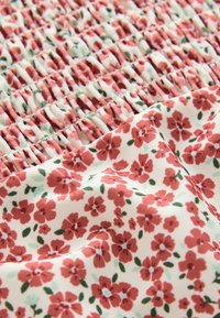 Tissu fleuri présentant de petites fleurs rouges sur un fond crème avec des feuilles vertes ; la texture froncée crée des sections rassemblées.