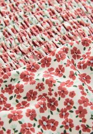 Tessuto floreale con piccoli fiori rossi su sfondo crema con foglie verdi; la trama arricciata crea sezioni raccolte.