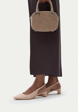 Osoba nosząca beżowe zamszowe czółenka na pasku i ciemnobrązową spódnicę, trzymająca pasującą beżową zamszową torebkę z logo marki.