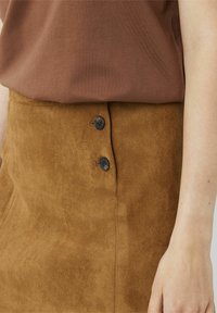 Jupe en daim marron avec deux boutons noirs sur le côté, à la texture lisse et à la coupe ajustée, partiellement couverte par un haut marron.