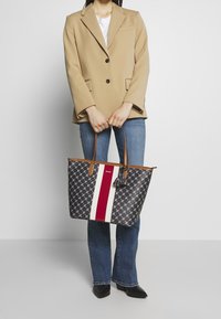 Nákupní taška s tmavým vzorovaným vnějškem, červeným a bílým pruhovým detailem a hnědými koženými držadly. Obsahuje dekorativní střapec.