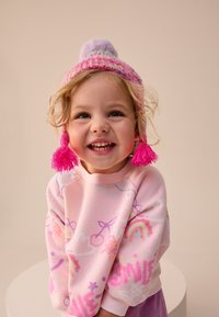 Rosa Sweatshirt mit bunten Mustern, lila und pinke gestrickte Mütze mit Bommel und pinke Quasten, lächelndes Kind, heller Hintergrund.