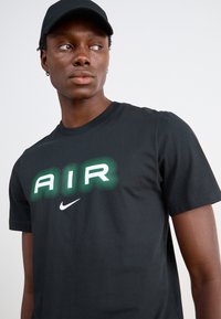Juodas medvilninis marškinėlis su žaliu ir baltu "AIR" grafiniu dizainu, kuriame yra bangos linijos, ir mažas baltas Nike logotipas žemiau. Užsidėta juoda kepurė.