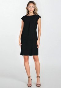 Czarna sukienka do kolan z krótkimi rękawami, z przodu z plisą. Tkanina ma gładką teksturę. Nosi się ją z wzorzystymi sandałami na wysokim obcasie.