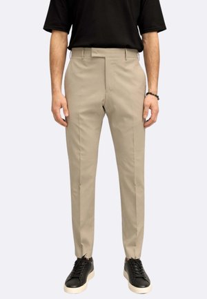 Uomo che indossa pantaloni beige slim fit, maglietta nera, sneakers nere con suole bianche, in piedi davanti a uno sfondo chiaro e semplice.