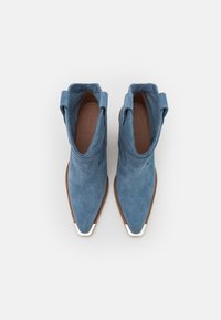 Botines de ante azul con punta afilada, tira de agarre redonda y detalles en tono plateado en la puntera. Suela plana de cuero y acabado texturizado.