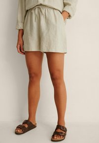 Linen shorts in light green with an elastic waistband and side pockets. Paired with brown slide sandals featuring two adjustable straps.