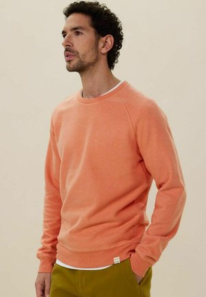 Sweatshirt corail en mélange de coton doux, avec des manches raglan, un col rond, des poignets et un ourlet côtelés. Porté par-dessus un t-shirt blanc.