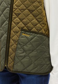 Γιλέκο με καπιτονέ ραφή σε ελιά και σκούρο πράσινο με κλείσιμο φερμουάρ και μια τσέπη. Φέρει το λογότυπο της Barbour με κίτρινο χρώμα στο περίγραμμα της τσέπης.