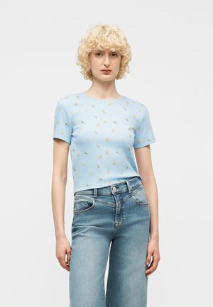 Ung person med kort, krøllete blondt hår som har på seg en lyseblå kortermet skjorte med små oransje frukttrykk og lyseblå jeans.