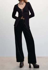 Czarny sweter z dzianiny z sznurkiem z przodu, zestawiony z czarnymi welurowymi spodniami o szerokich nogawkach i platformowymi butami na chunky obcasie. Widoczne szczegóły teksturalne.