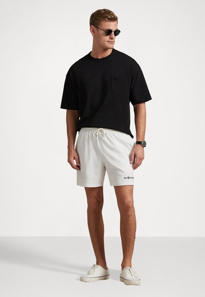 Juodas trumpas rankovės marškinėlis su krūtinės kišenėle, derinamas su baltais šortais su raišteliu ir mažu juodu logotipu. Baltos sportbačiai.