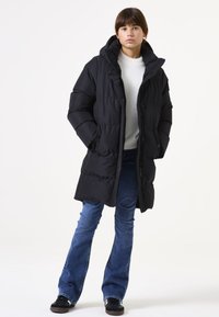 Abrigo de plumas negro con capucha, con un diseño acolchado y cremallera frontal. Combinado con vaqueros azules de pernera acampanada y zapatos negros.