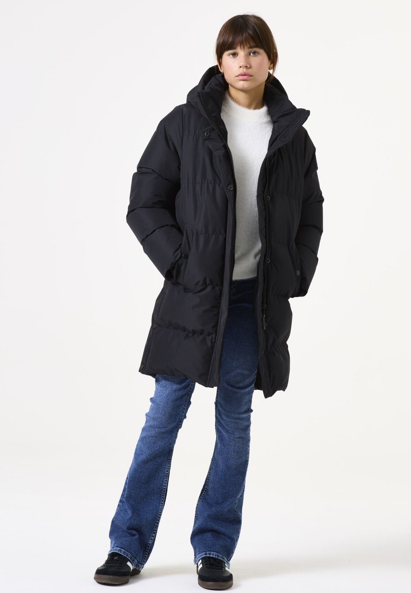 Abrigo de plumas negro con capucha, con un diseño acolchado y cremallera frontal. Combinado con vaqueros azules de pernera acampanada y zapatos negros.