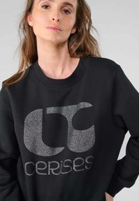 Sweatshirt noir à encolure ronde, avec un grand logo argenté brillant et du texte sur le devant. Fabriqué en tissu doux avec une coupe décontractée.
