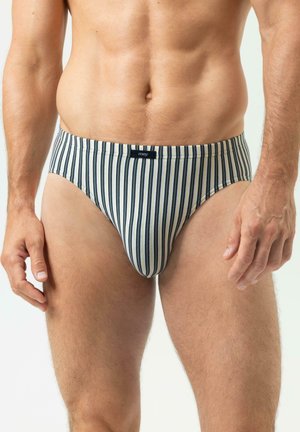 Mann trägt weiße Unterhose mit vertikalen schwarzen und grauen Streifen, zeigt Oberkörper und obere Beine vor einem einfachen weißen Hintergrund.