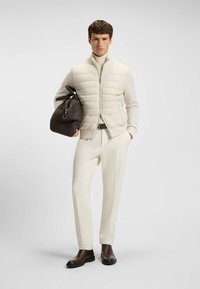 Leichte, cremefarbene Puffer-Weste, eng anliegender weißer Rollkragenpullover, maßgeschneiderte weiße Hose, braune Ledertasche und braune Stiefeletten mit schwarzen Sohlen.