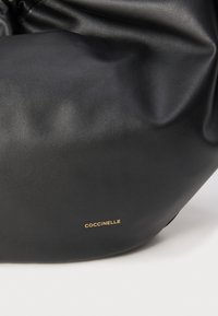 Zachte zwarte leren handtas met subtiele textuur, gerimpeld ontwerp en klein gouden "Coccinelle"-logo aan de onderkant van de voorkant.