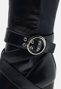 Bota de cuero azul marino hasta la rodilla con un acabado brillante, que presenta una correa en forma de cruz con una hebilla redonda plateada y ojetes.