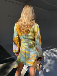 En kvinne med bølgete blondt hår har på seg en fargerik, mønstret kjole og holder en perleveske, i et lyst, minimalistisk studio.