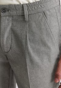 Nahaufnahme von strukturierten grauen Hosen mit Knopf und Gürtelschlaufe, die eine Hand zeigt, die an den Oberschenkel gelehnt ist.