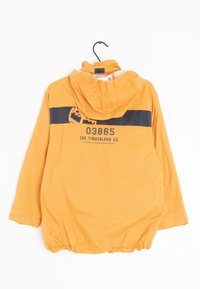 Senapsgul Timberland vindjacka med huva, svart horisontell rand och logotexten "03885 The Timberland Co." på baksidan, hängande på en svart krok.