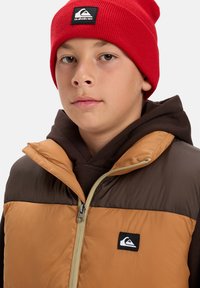 Ung person som bär en röd mössa, brun hoodie och en beige och mörkbrun puffväst med ett Quiksilver-logotypmärke.