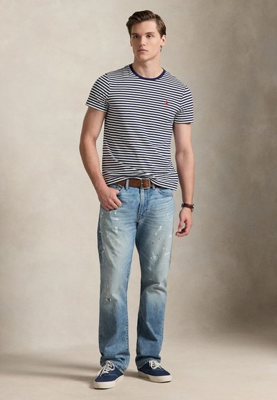 Νεαρός άνδρας στέκεται με το χέρι στην τσέπη, φορώντας μπλε-λευκή ριγωτή μπλούζα, ανοιχτό μπλε τζιν με πιτσιλιές μπογιάς, καφέ ζώνη και μπλε αθλητικά παπούτσια.