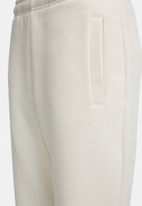 Ljuscremefärgade joggerbyxor gjorda av mjukt tyg, med en platt framficka och präglad logotyp på sidan. En enkel, skräddarsydd design.