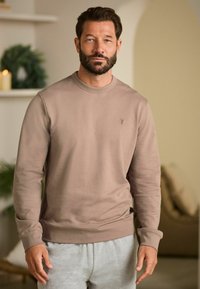 Sweatshirt beige en tissu doux avec des manches longues et un col rond. Présente un petit logo sur la poitrine. Assorti avec un pantalon de survêtement gris.
