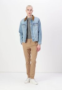 Light blue denim jacket with brown collar, gray shirt, khaki pants, and white sneakers. Model standing on white background.