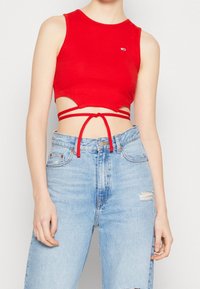 Mujer con un top corto rojo con lazos alrededor y unos jeans de tiro alto y rotos de color azul claro, de pie contra un fondo liso.
