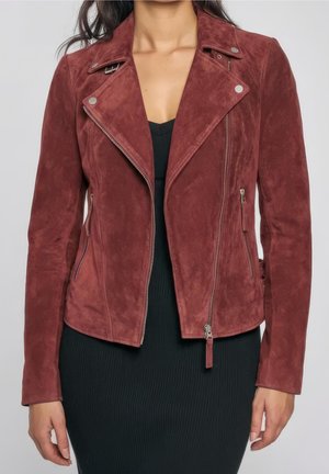 Femme portant une veste de motard ajustée en daim couleur rouille avec des fermetures éclair argentées, par-dessus une robe noire côtelée, visible du cou jusqu'aux hanches.