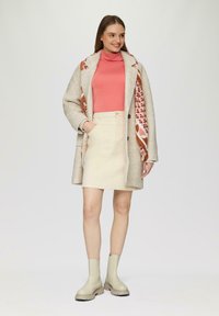 Beige Wollmantel mit großen Knöpfen, kombiniert mit einem rosa Rollkragenpullover, einem cremfarbenen Rock und hellgrauen Ankle Boots. Ein gemusterter Schal fügt Farbe hinzu.