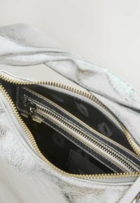 Strieborná metalická taška s textúrovaným povrchom, so zipsovým uzáverom, zlatým zipsovým detailom a viacerými čiernymi vnútornými vreckami.