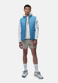 Veste matelassée bleu clair sur un sweat à capuche blanc, accompagnée de shorts beiges et de baskets texturées. Des chaussettes blanches avec un design noir.